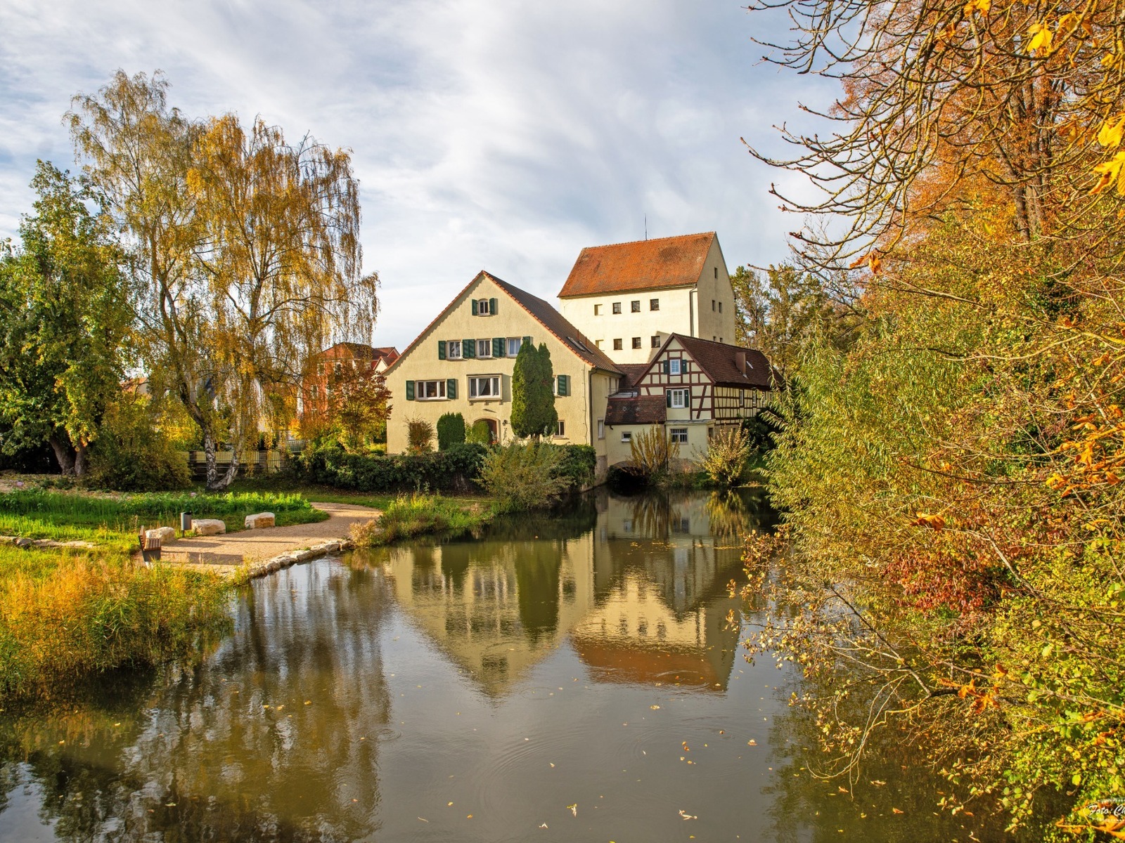 Die Feuchtwanger Stadtmühle und ein Fachwerkhaus spiegeln sich in einem Gewässer, umgeben von Herbstbäumen unter blauem