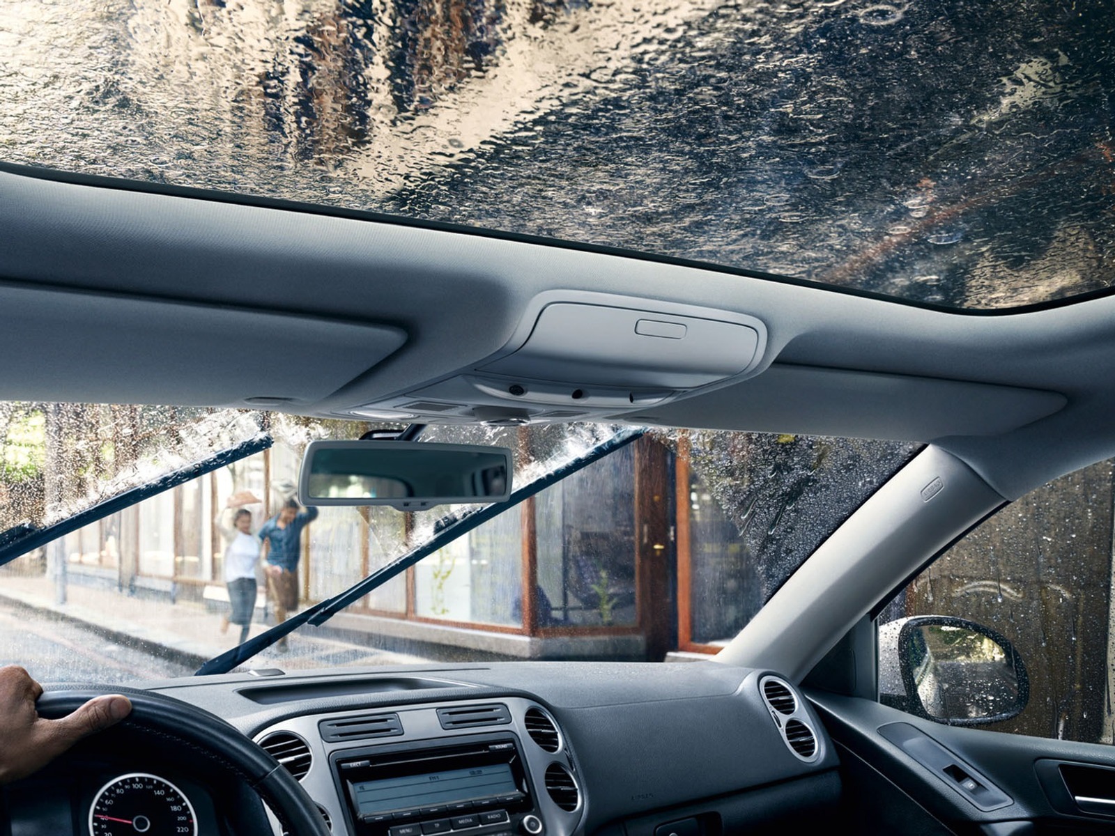 Eine Hand am Lenkrad eines Autos, dessen Scheibenwischer Regen von der Windschutzscheibe wischen und ein nasses