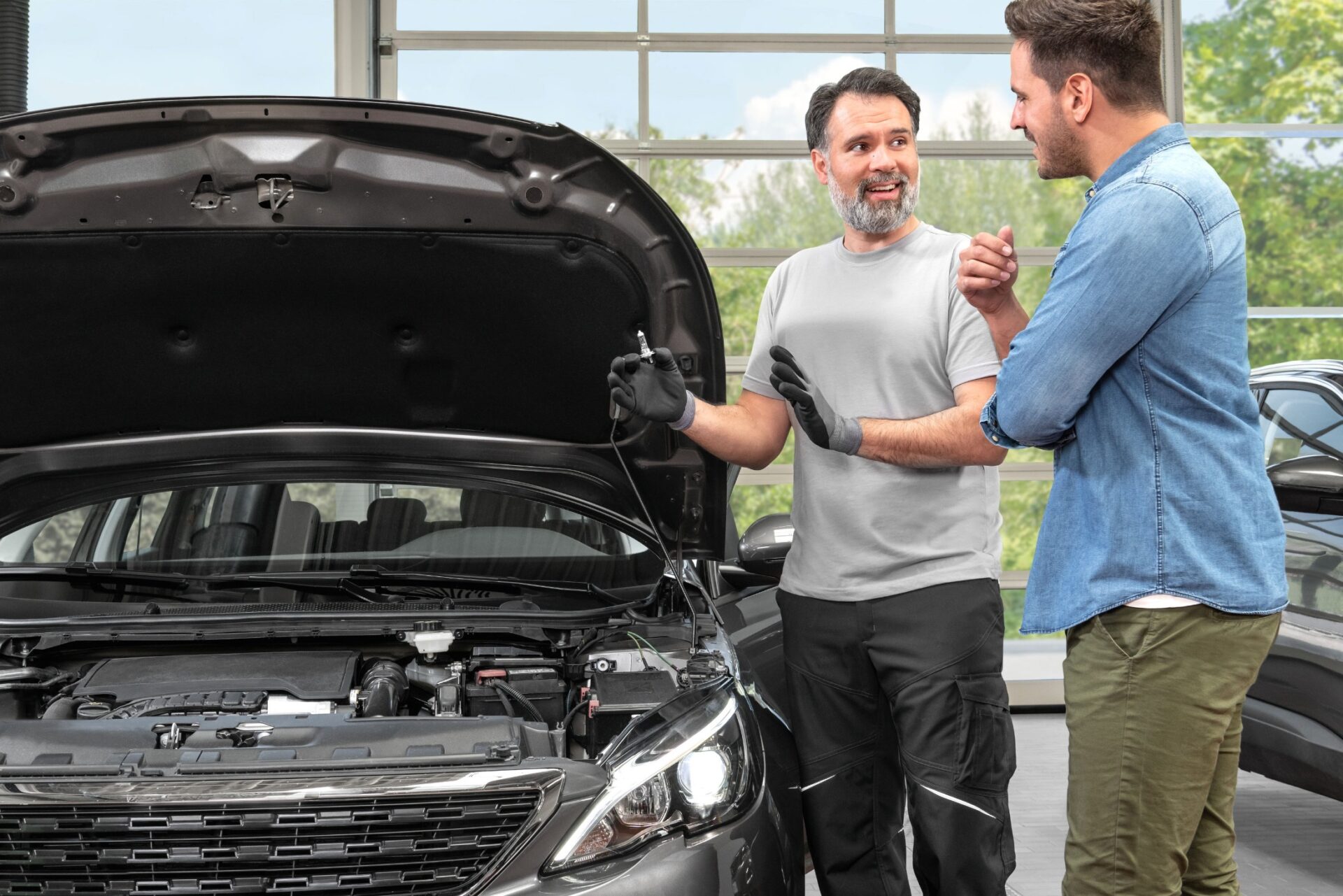 Mechaniker zeigt einem Kunden in einer Werkstatt eine Glühbirne am geöffneten Motorraum eines Autos