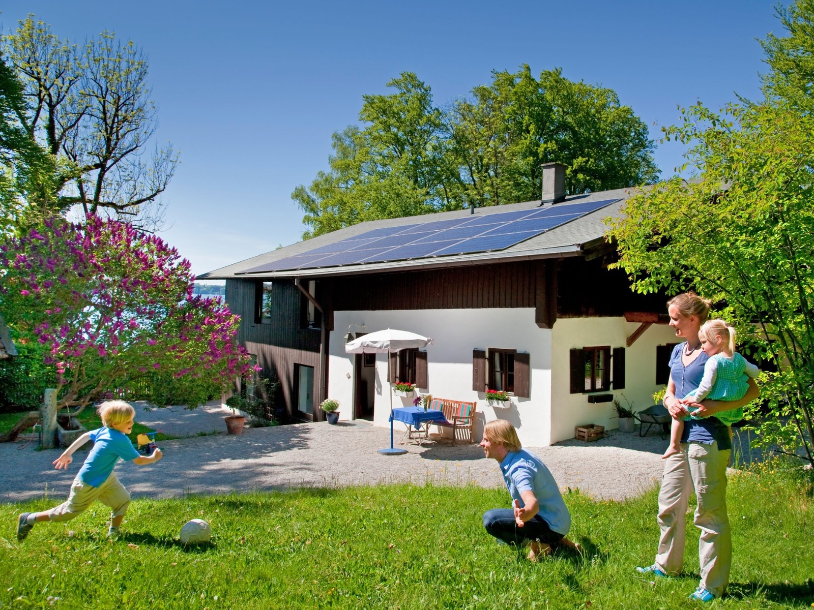 Eine Familie spielt im Garten vor einem Haus mit Solaranlage, ein Kind rennt auf einen Ball zu, Frau hält Kleinkind