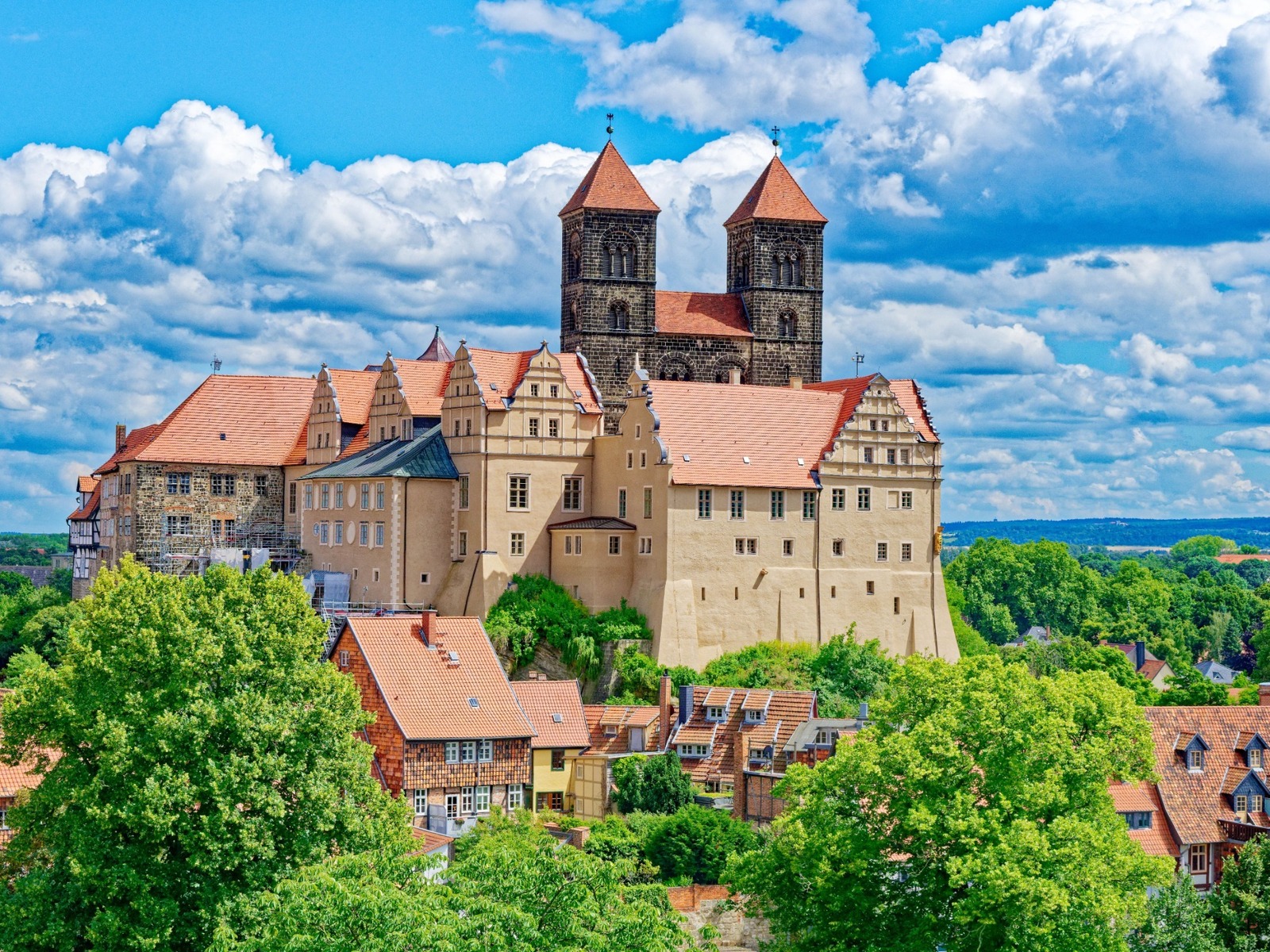 Historische Burganlage mit zwei hohen Türmen und roten Dächern, umgeben von grünen Bäumen unter blauem Himmel mit Wolken