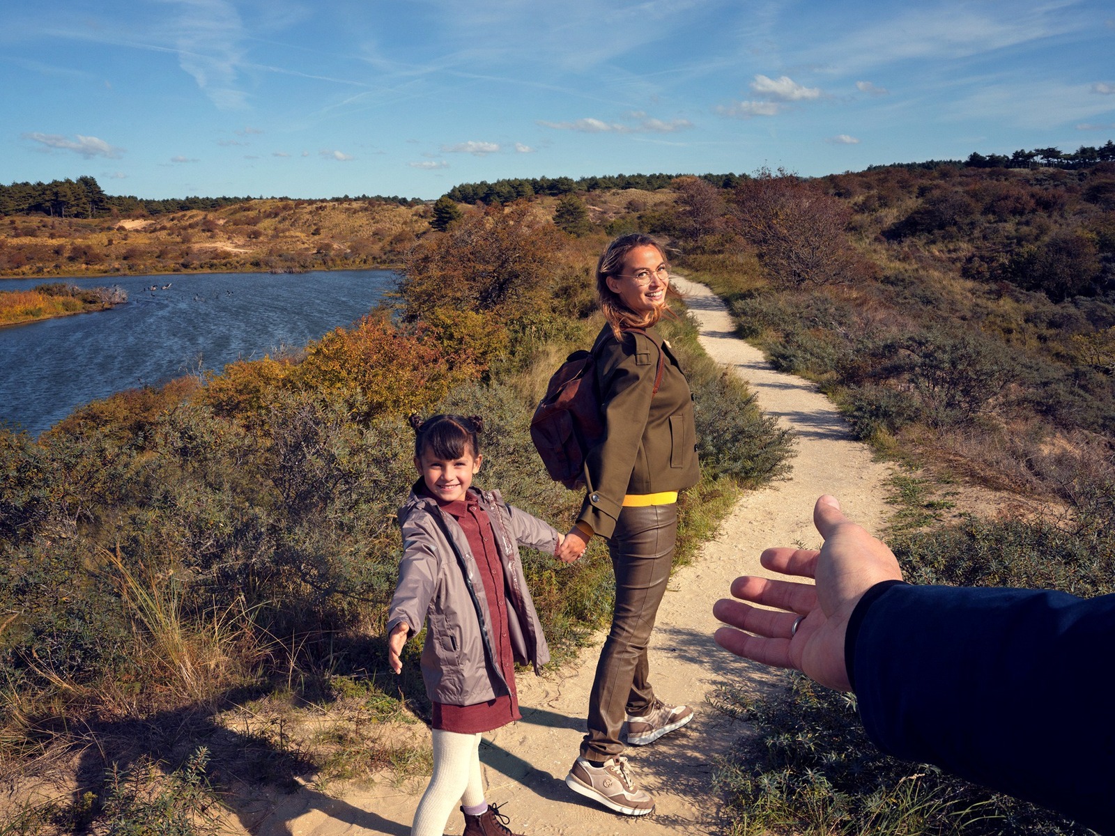 Frau und Kind wandern lächelnd auf einem Pfad entlang eines Sees, halten Händchen, eine Hand ist im Vordergrund