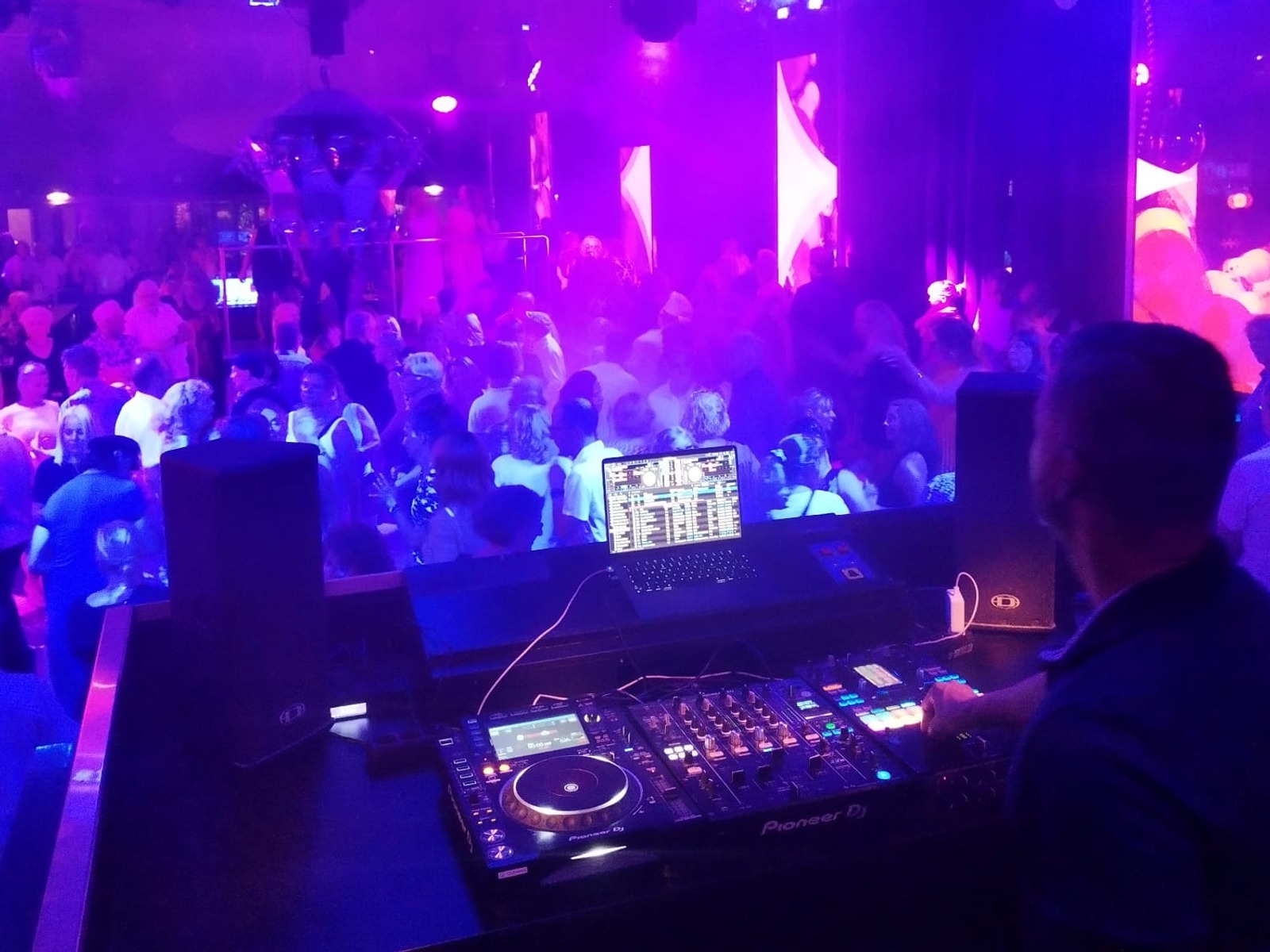 DJ an einem Mischpult legt Musik auf, während viele Menschen auf einer Tanzfläche unter blau-violettem Licht tanzen