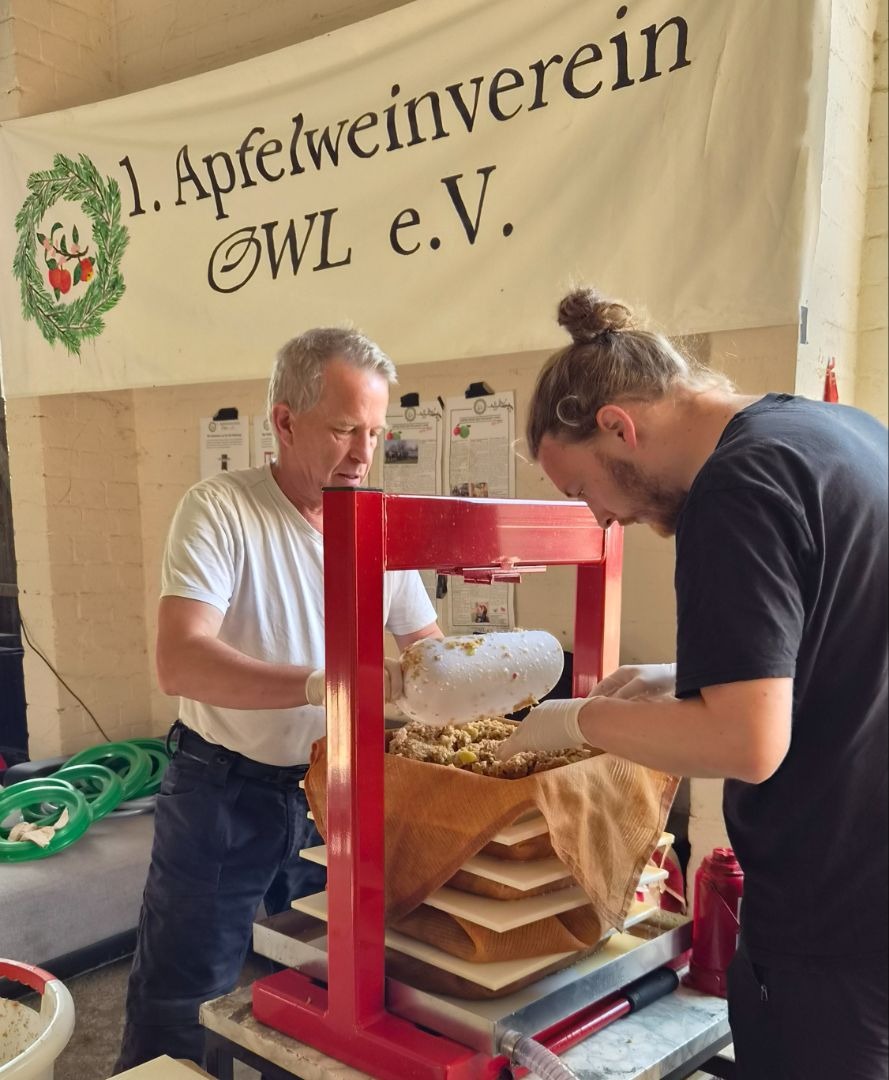 Zwei Männer bedienen eine rote Apfelpresse, einer füllt Maische ein, vor dem Banner des 1. Apfelweinverein OWL e.V