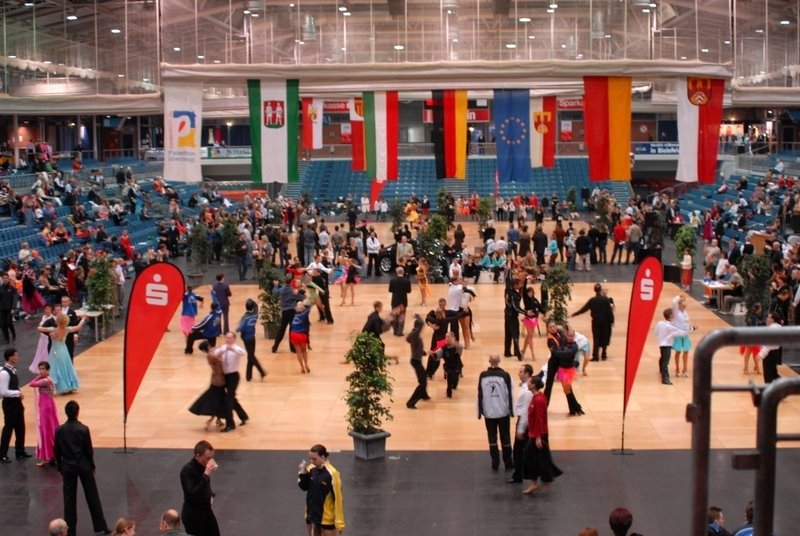 Tanzpaare in festlicher Kleidung tanzen auf hellem Parkett in einer großen Halle mit Zuschauern und Nationalflaggen