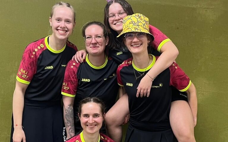Fünf lachende Frauen im Team-Sportoutfit posieren, eine huckepack auf einer anderen, eine kniend davor