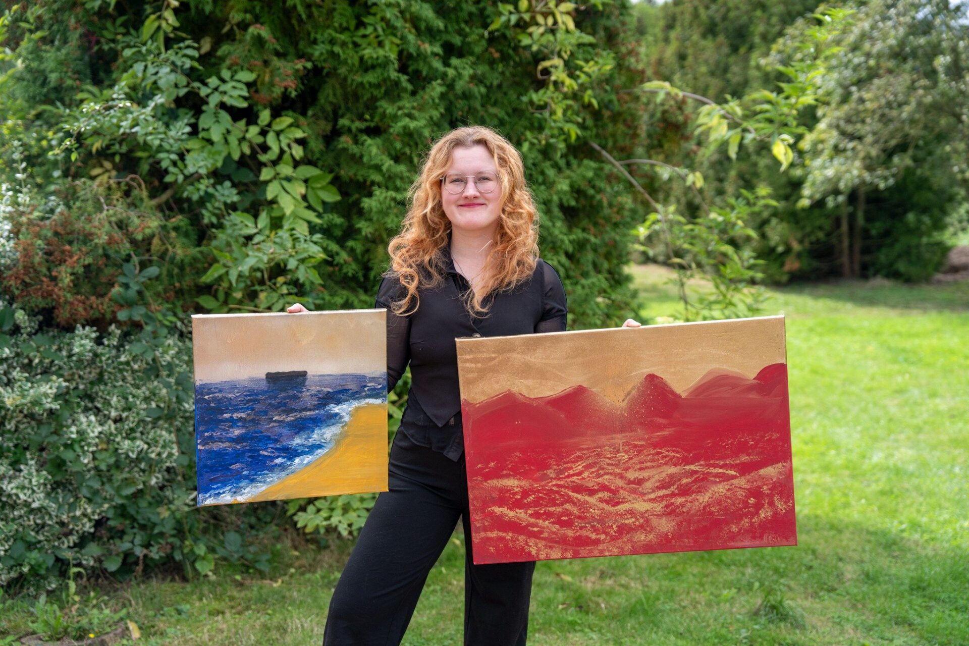 Michelle Roth hält zwei Gemälde: eines mit Meer und Sand, ein anderes abstrakt in Rot-Gold vor grüner Natur