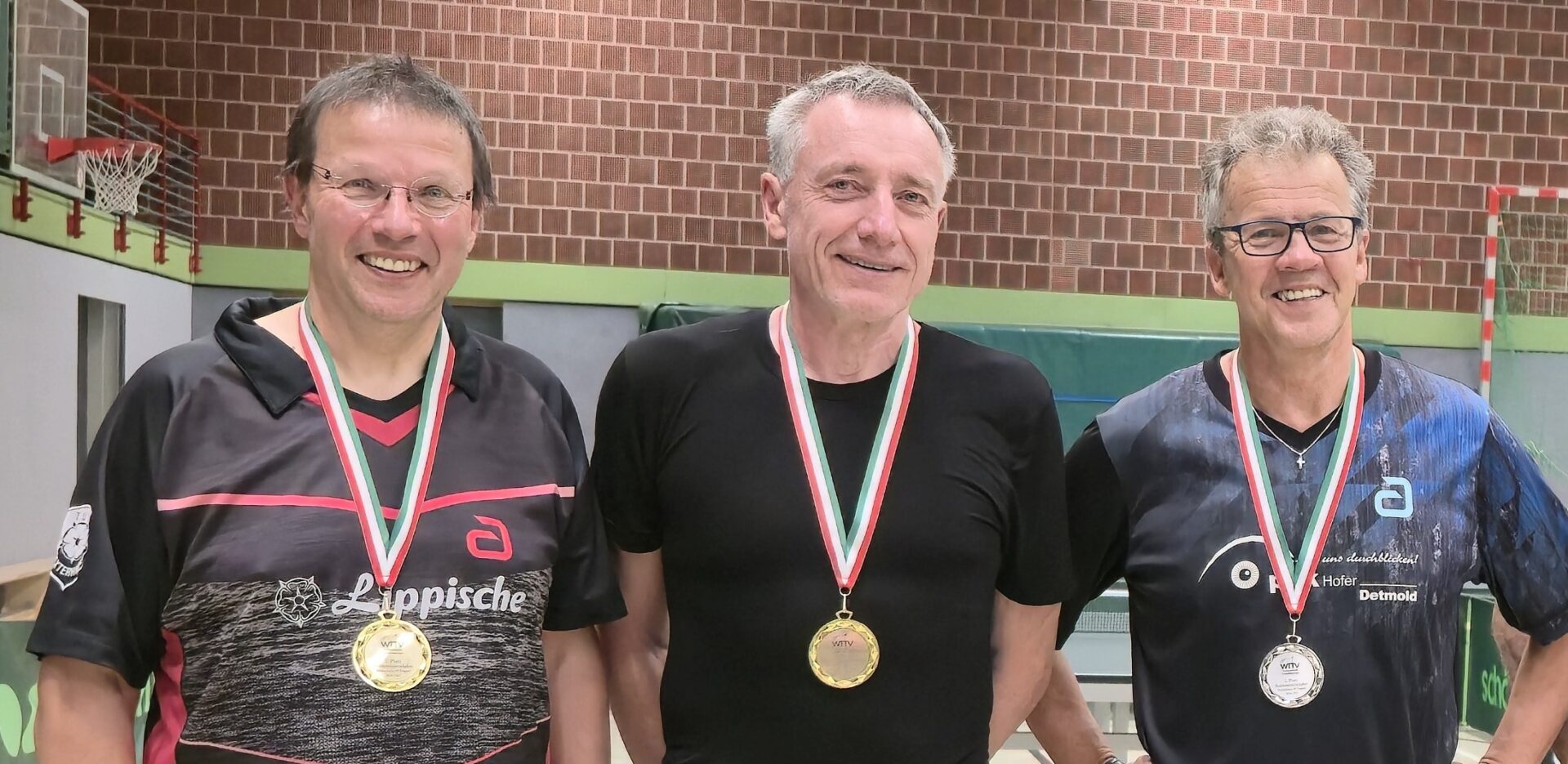 Drei lächelnde Männer mit Medaillen um den Hals in einer Sporthalle