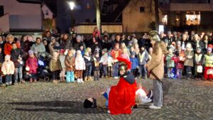 Viele Zuschauer mit Laternen sehen zu, wie Sankt Martin seinen Mantel mit einem Bettler teilt