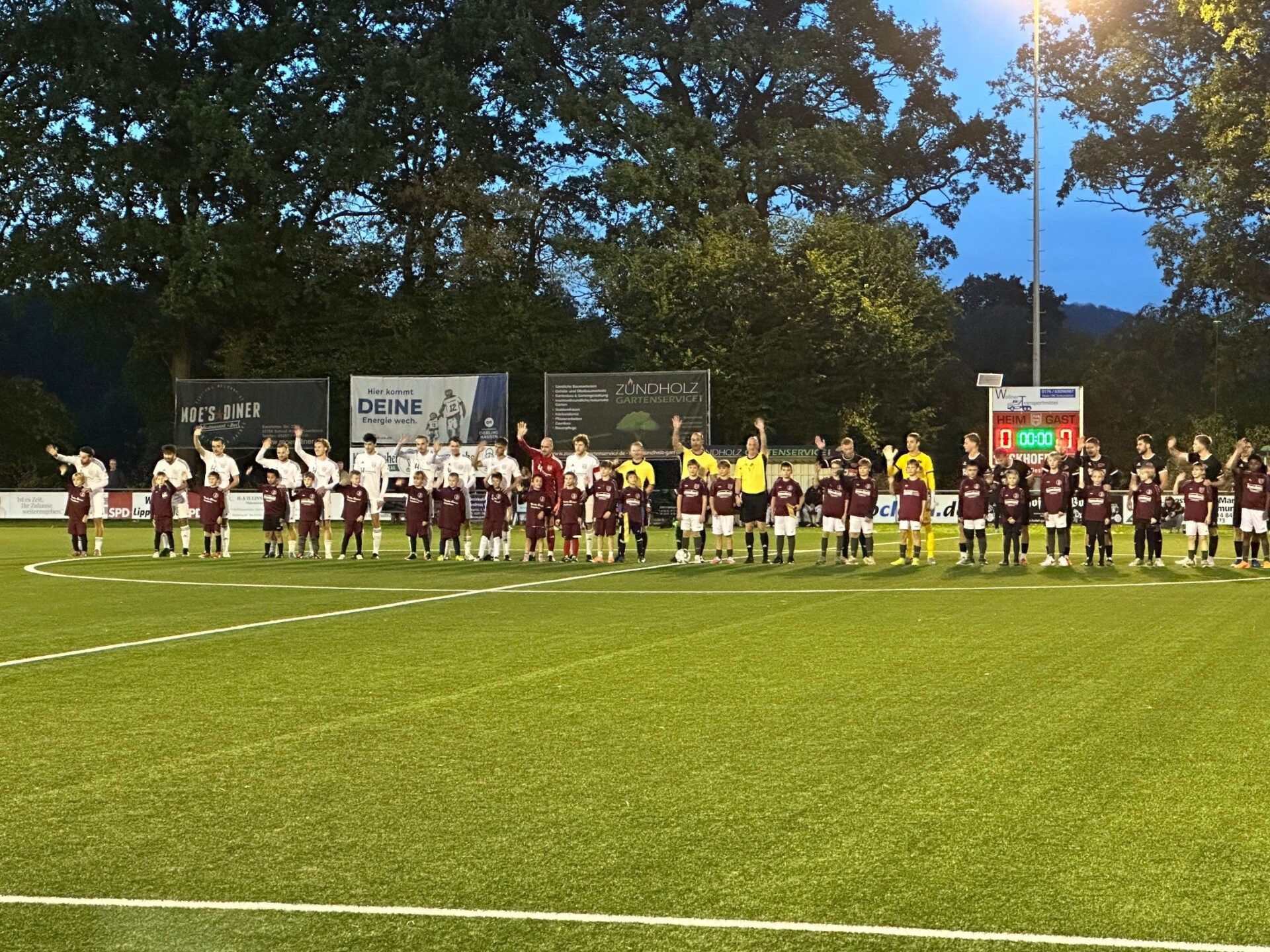 Zwei Fussballmannschaften mit Schiedsrichtern und Kindermaskottchen stehen auf einem Flutlicht-Spielfeld und winken