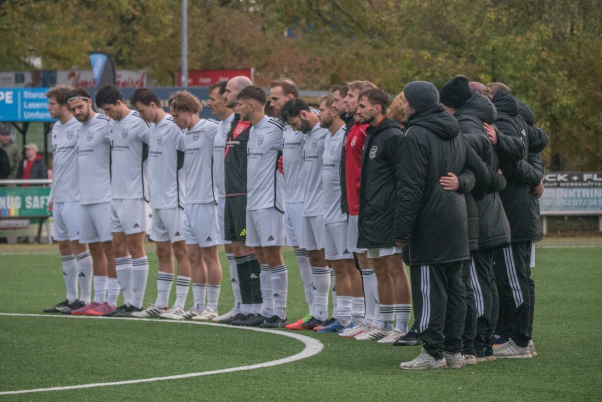Eine Reihe von Fußballspielern und Trainern in weißen Trikots und dunklen Jacken steht eng auf einem Rasenplatz