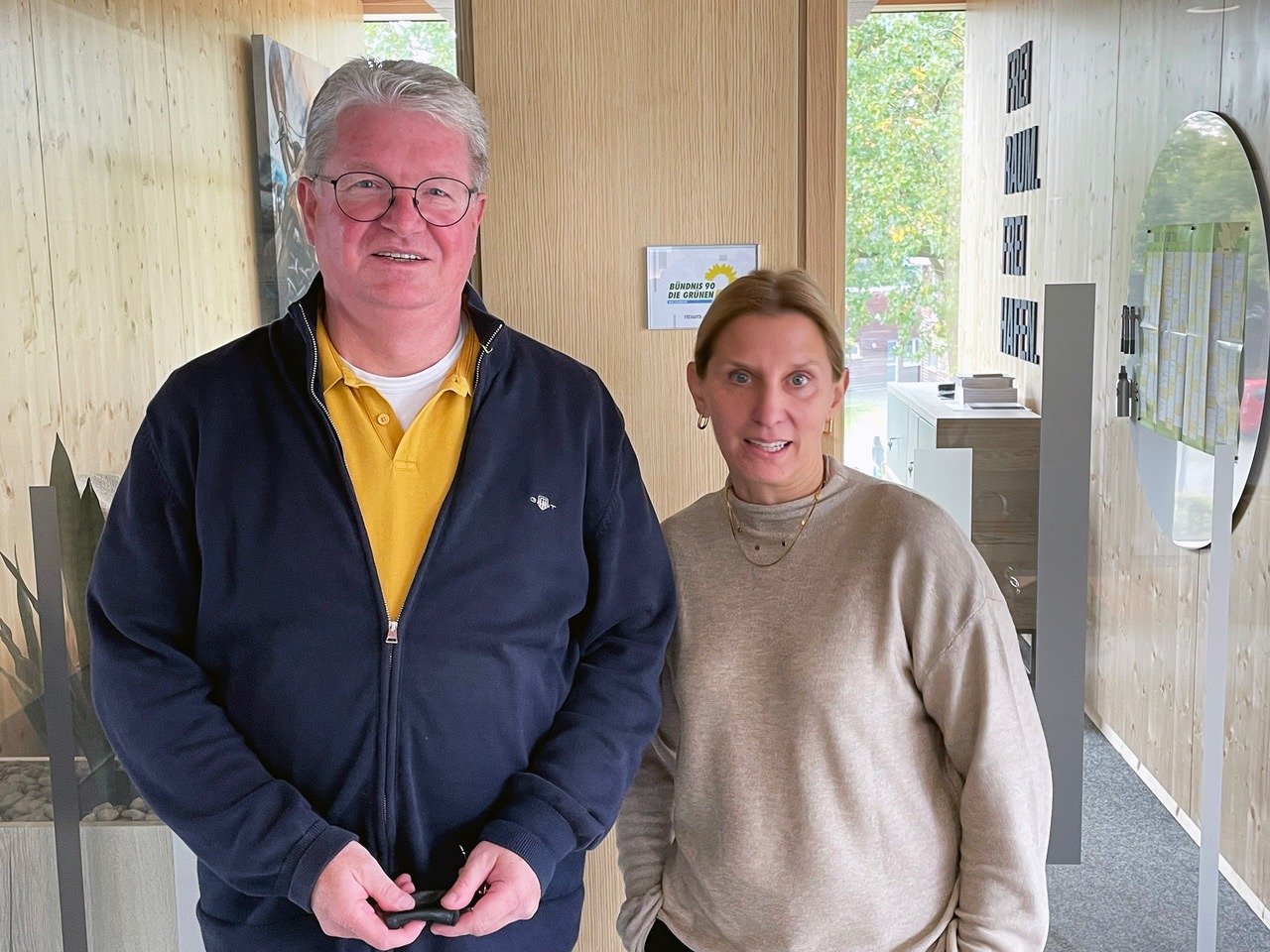 Mann und Frau in einem Büro vor einer Holzwand mit dem Logo von Bündnis 90 Die Grünen