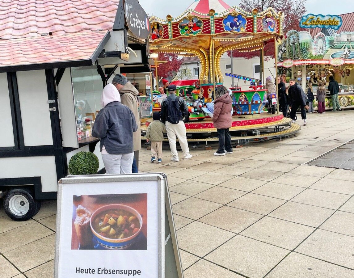 Besucher warten an einem Crêpes-Stand und stehen um ein buntes Karussell auf dem Herbstkirmes-Marktplatz