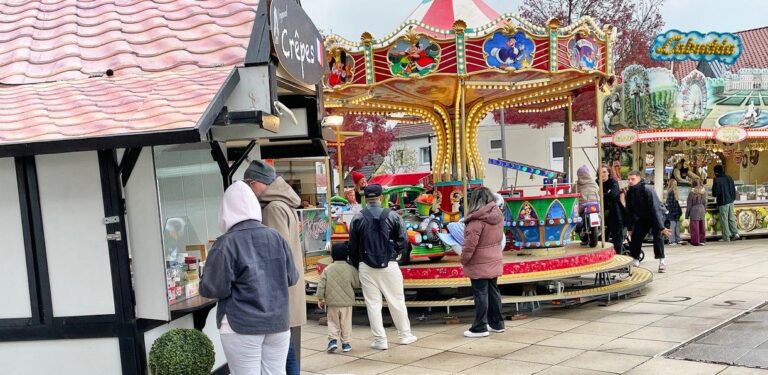 Besucher warten an einem Crêpes-Stand und stehen um ein buntes Karussell auf dem Herbstkirmes-Marktplatz