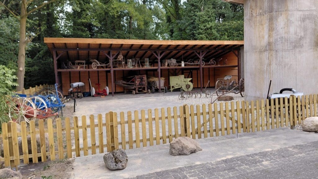 Offener Holzschuppen mit alten Landmaschinen, daneben ein Betonsilo, umgeben von Bäumen, davor ein Holzzaun