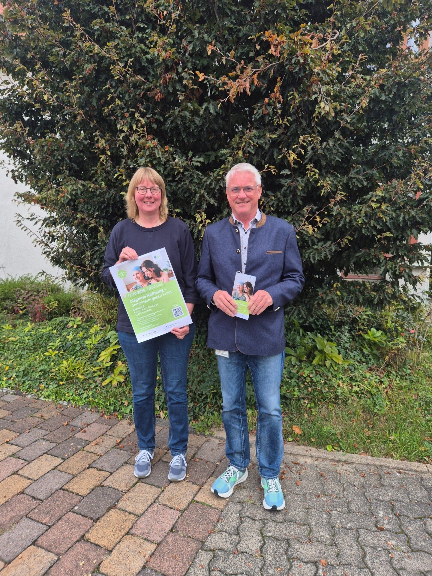 Zwei lächelnde Personen, ein Mann und eine Frau, halten Informationsflyer vor einem grünen Busch und einem Pflasterweg