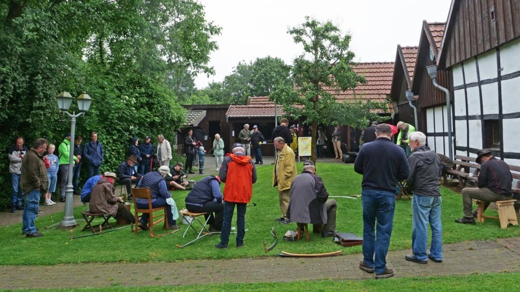 Eine Gruppe Menschen versammelt sich auf einer grünen Wiese, einige sitzen, vor traditionellen Gebäuden