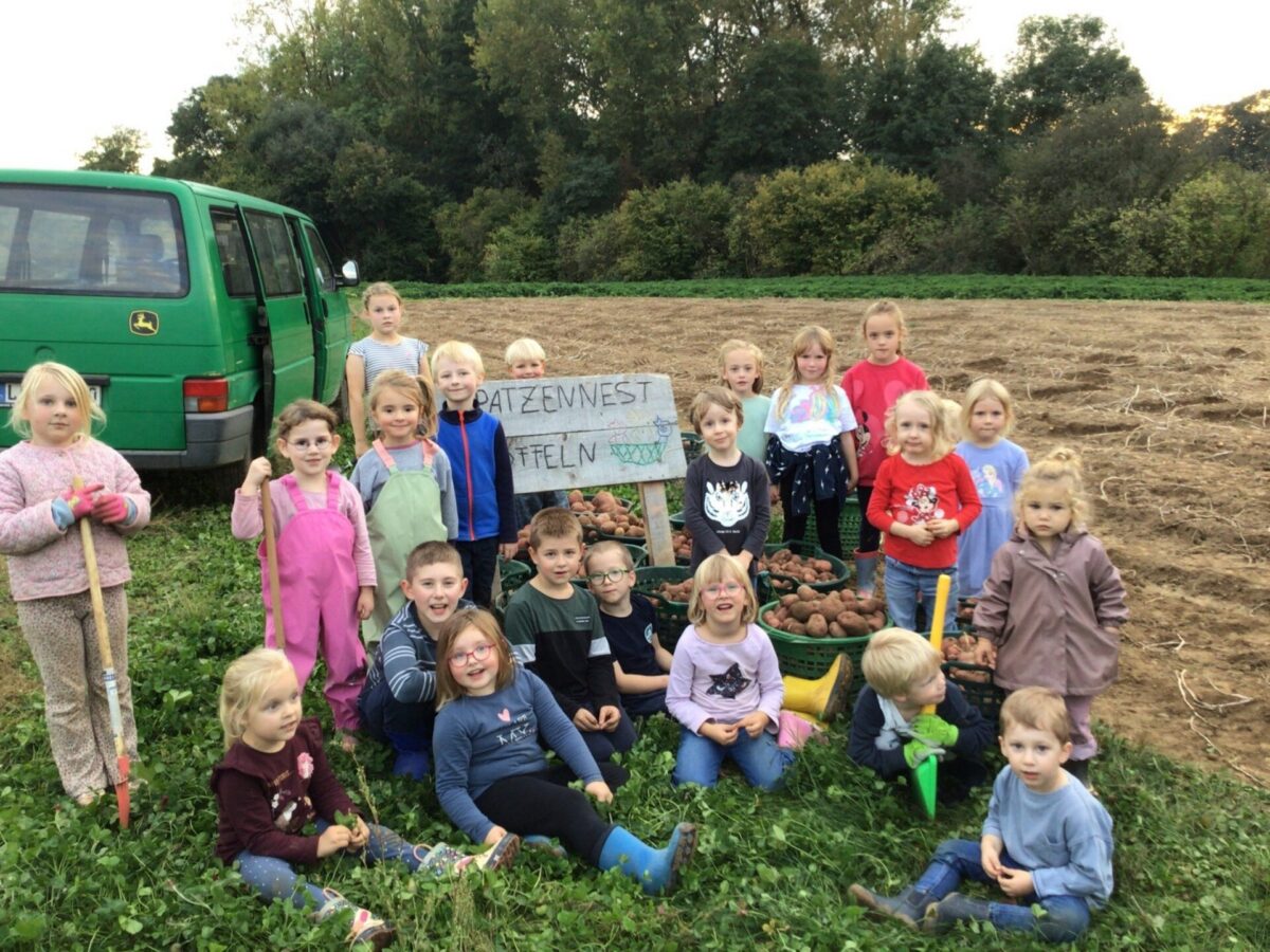 Eine Gruppe Kinder steht und sitzt in einem Kartoffelfeld vor gefüllten Erntekörben und einem grünen Bus