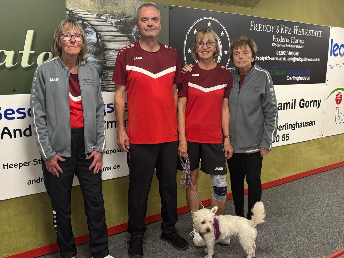Zwei Männer und zwei Frauen in roten und grauen Sportoutfits stehen mit einem kleinen weißen Hund vor einer Sponsorwand