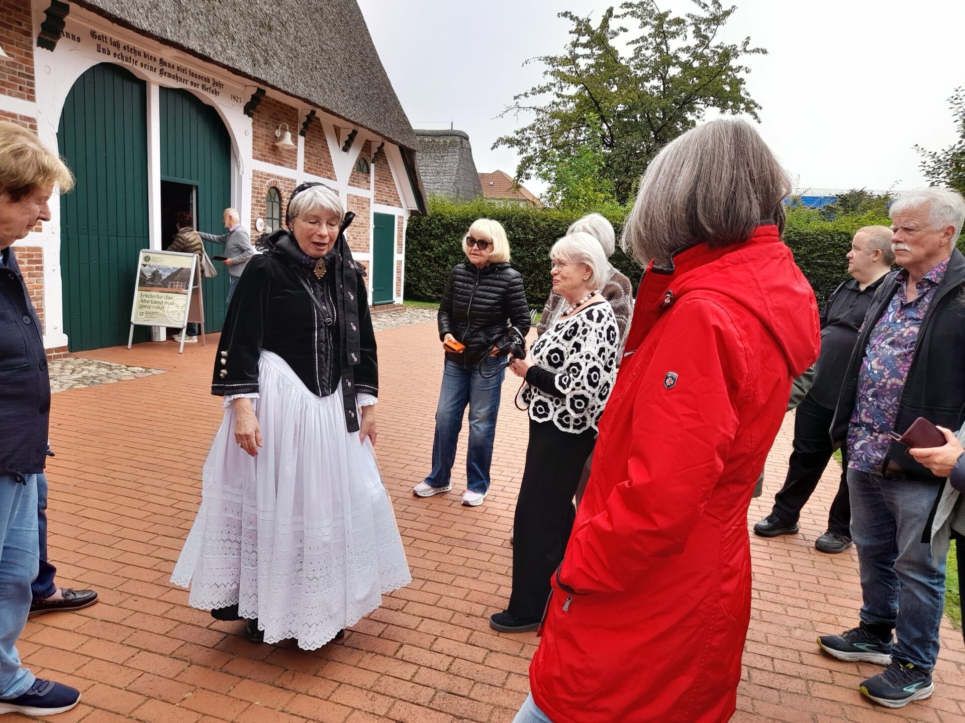 Mehrere Personen, eine Frau in schwarz-weißer Tracht, stehen auf einem Pflasterweg vor einem historischen Bauernhaus