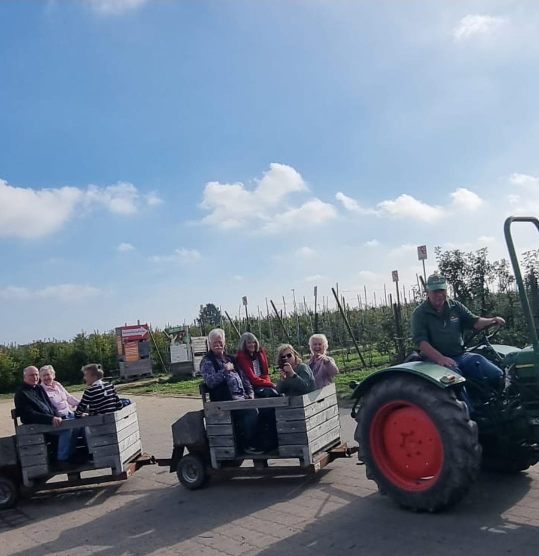 Ein Mann fährt einen grünen Traktor, der zwei Holzanhänger mit mehreren Senioren durch einen Obstgarten zieht