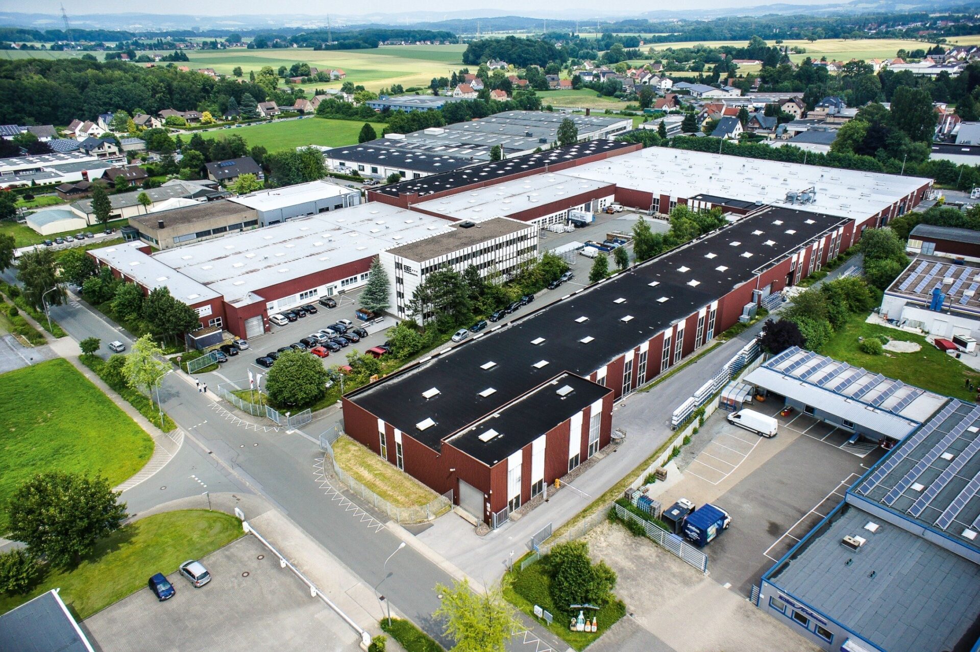 Luftaufnahme eines Industrieparks mit mehreren großen Hallen, Straßen, Parkplätzen und umliegenden Wohngebieten