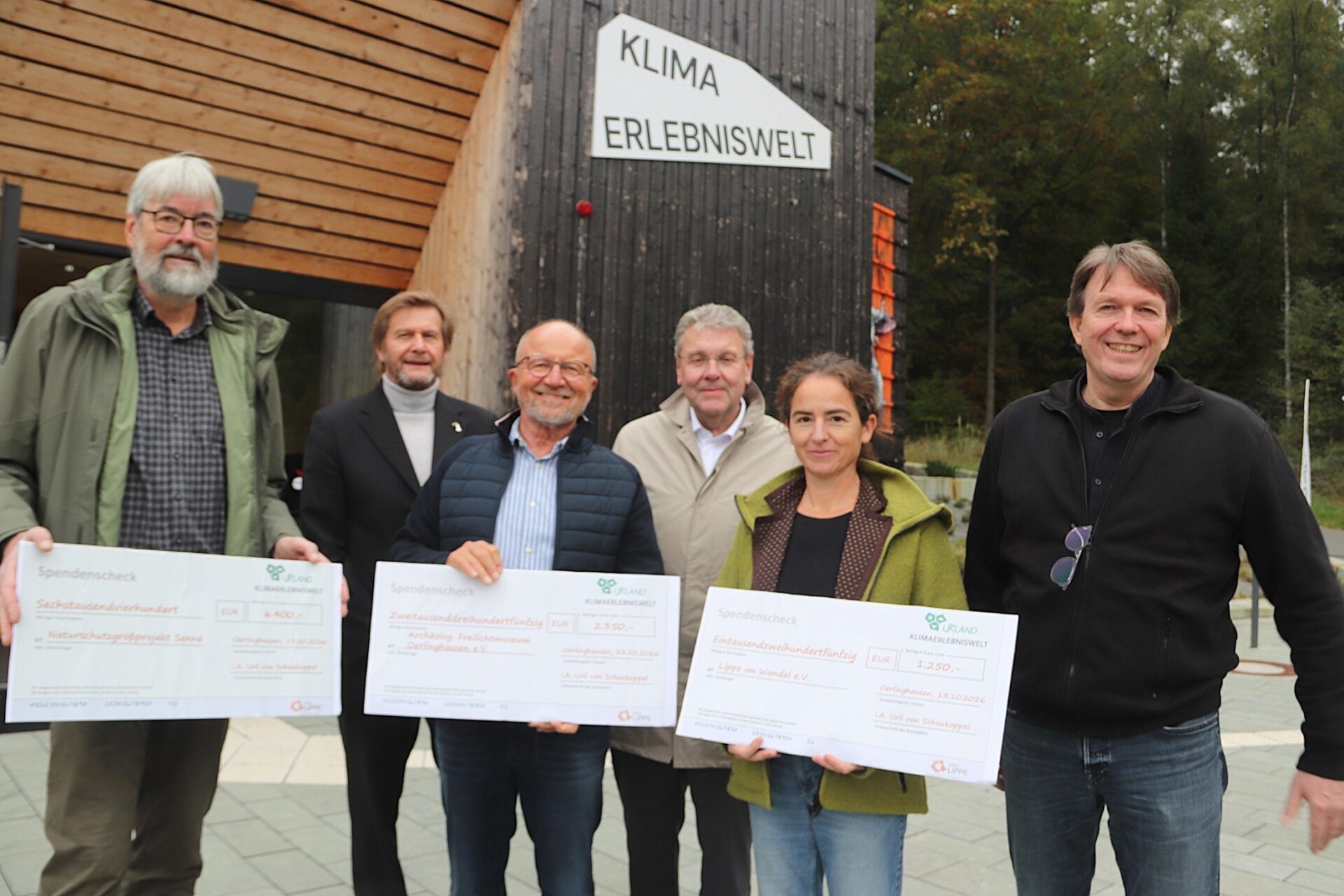 Sechs lächelnde Personen präsentieren drei Spendenschecks vor dem Gebäude der Klima Erlebniswelt