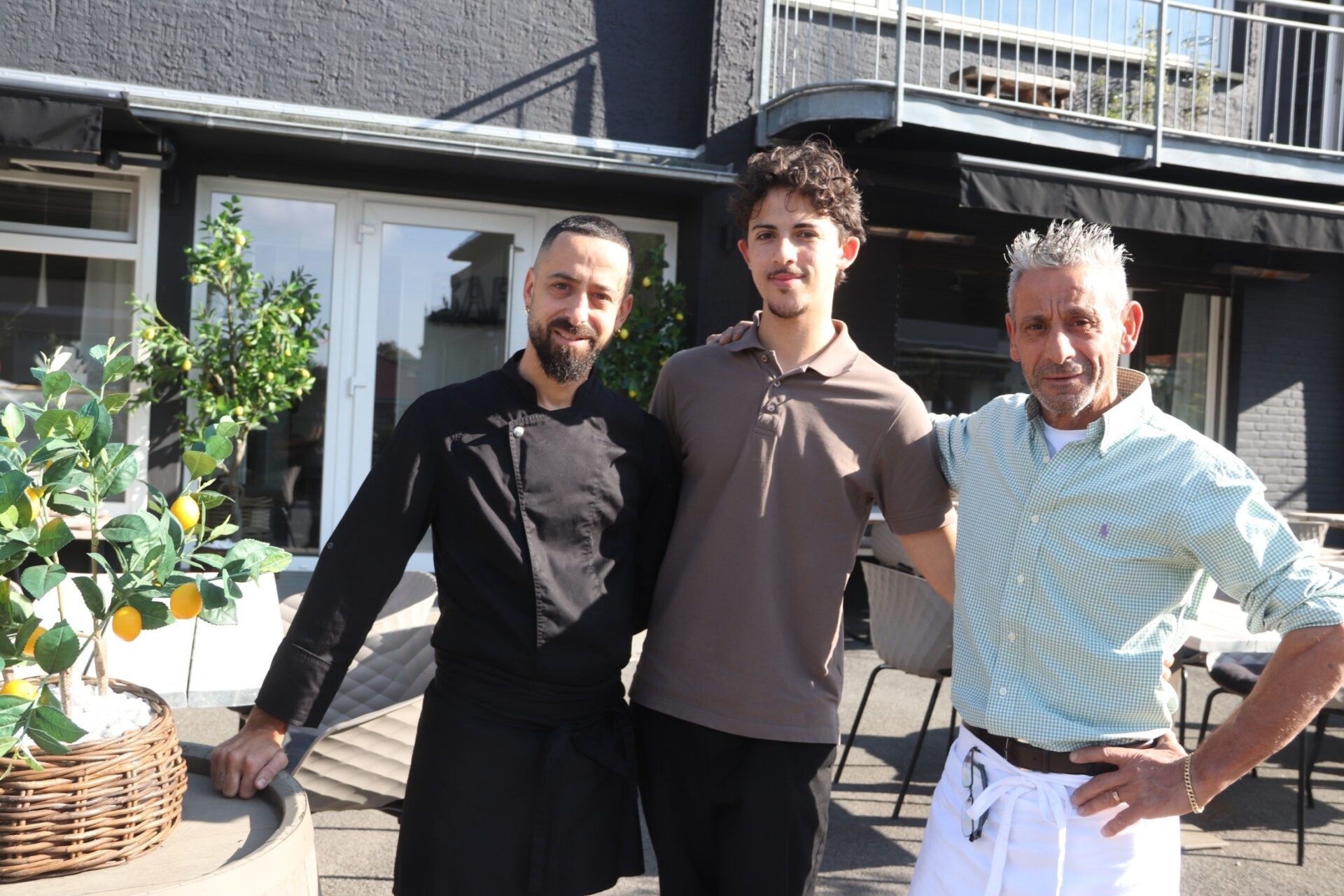 Drei Männer, zwei davon in Gastronomie-Kleidung, stehen lächelnd vor dem Eingang eines Restaurants mit Zitronenbäumen