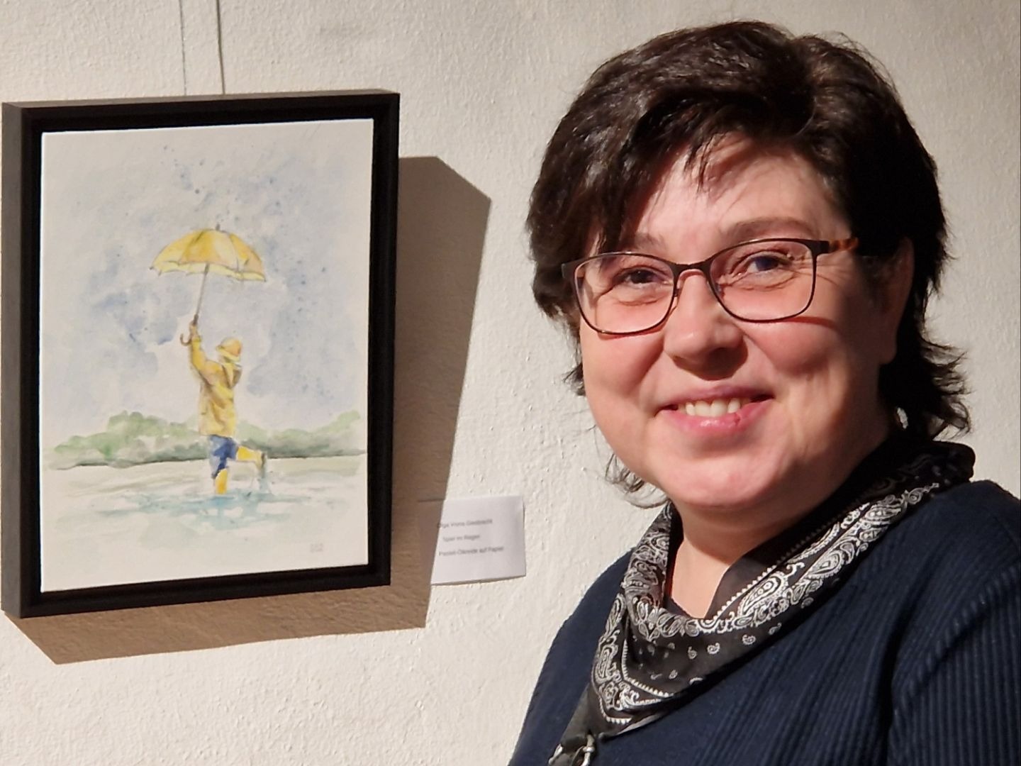 Künstlerin Olga Vons Giesbrecht lächelt neben ihrem Aquarell "Spiel im Regen" mit Kind und gelbem Schirm