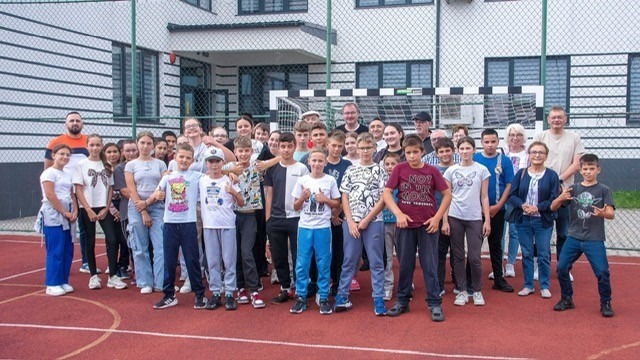 Große Gruppe Kinder, Jugendlicher und Erwachsener steht auf einem roten Sportplatz vor einer Schule mit Tor
