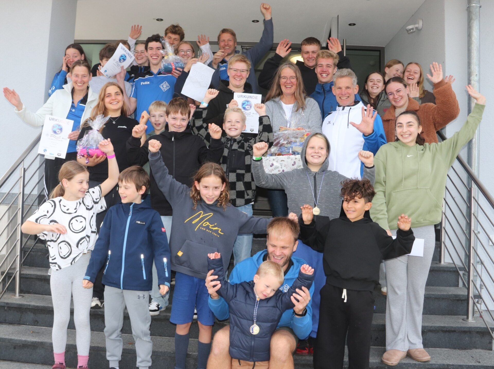 Eine große Gruppe lachender Kinder und Erwachsener steht auf einer Treppe und feiert mit Urkunden und erhobenen Armen