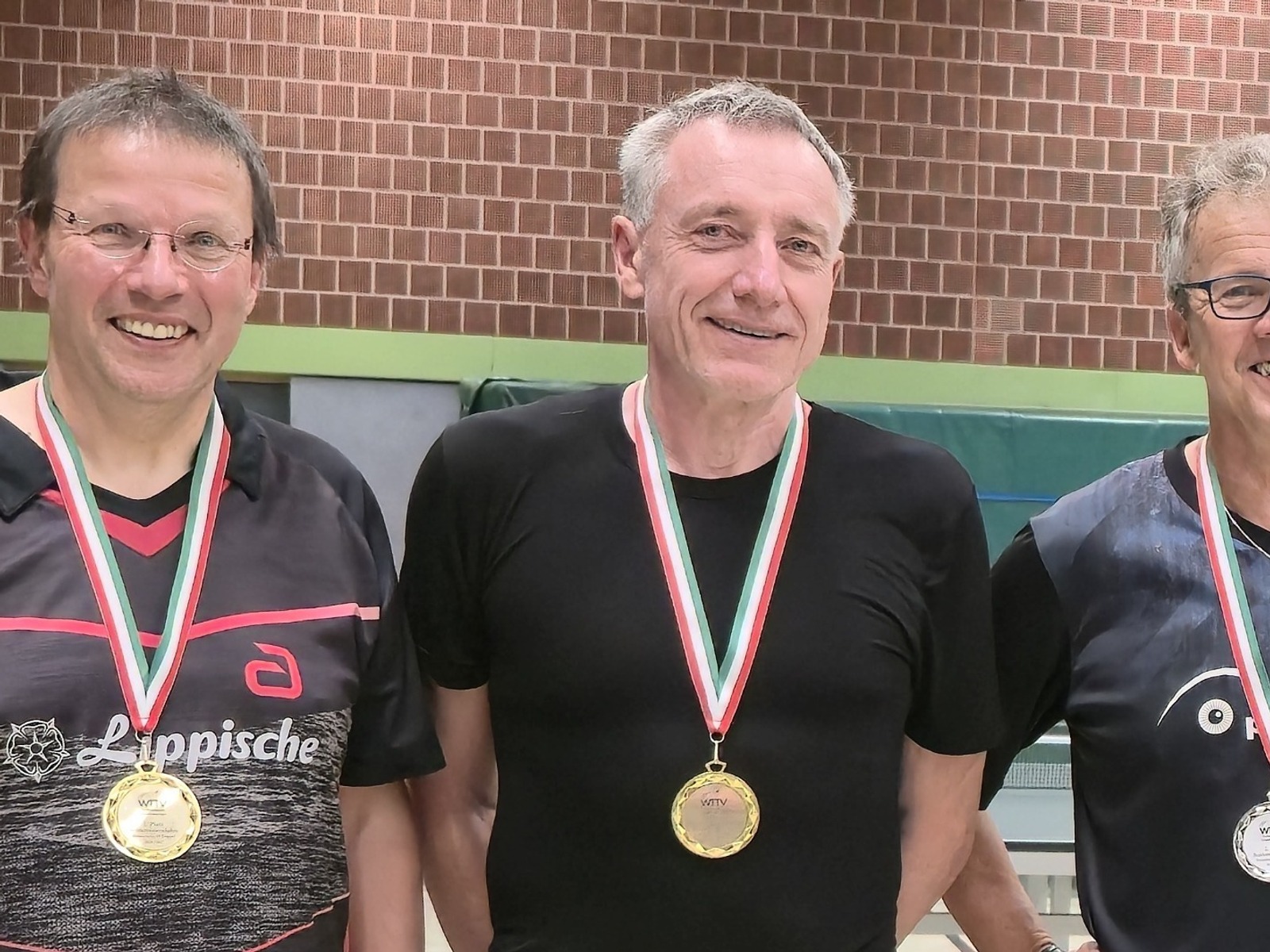 Drei lächelnde Männer mit Medaillen um den Hals in einer Sporthalle