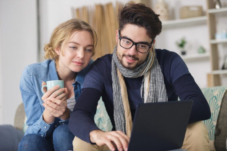 Ein Mann mit Brille und Schal tippt am Laptop, während eine Frau mit Tasse daneben zusieht