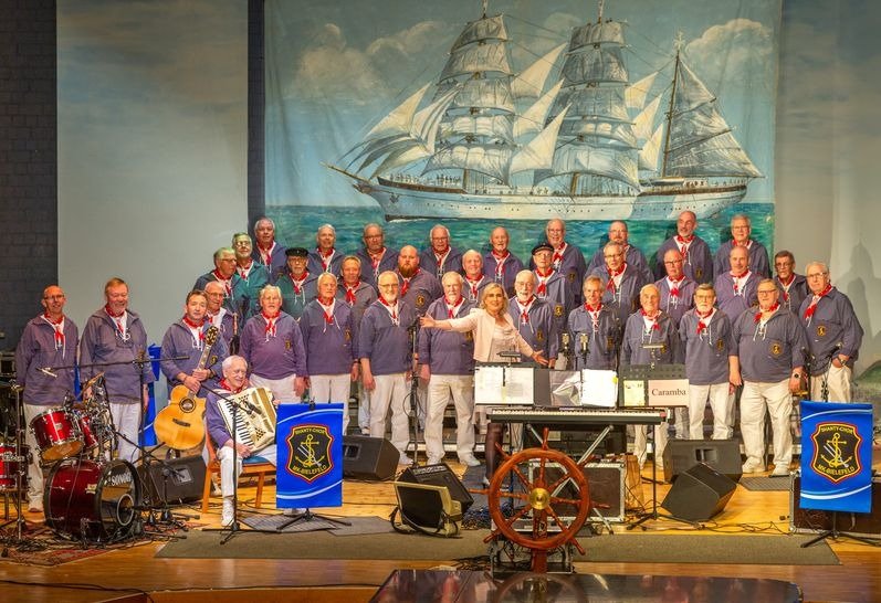 Großer Shanty-Chor mit einer Dirigentin auf der Bühne vor einem Segelschiff-Hintergrund, mit Instrumenten wie Akkordeon