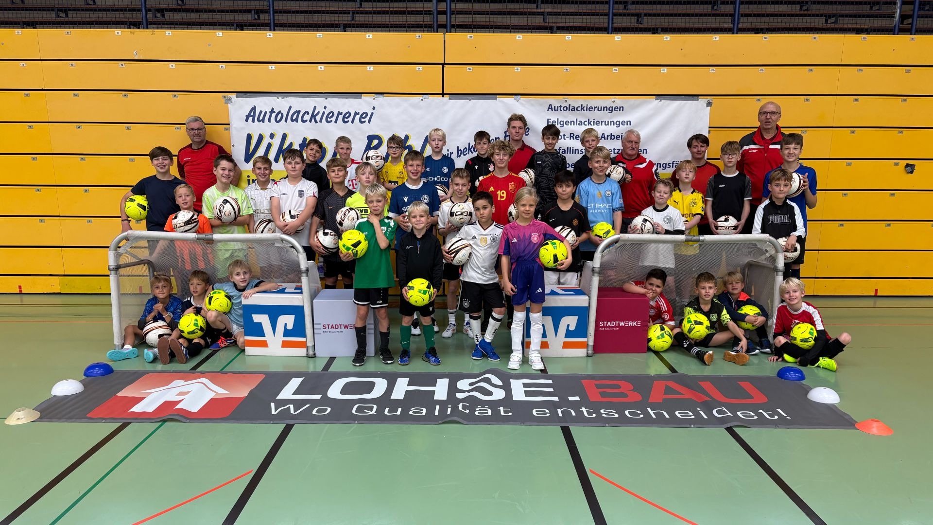 Viele lachende Kinder und erwachsene Trainer mit Fußbällen stehen und sitzen in einer Sporthalle