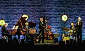 Fünf Musiker in Anzügen spielen Jazz auf einer Bühne mit Klavier, Gitarre, Kontrabass, Schlagzeug und Klarinette vor