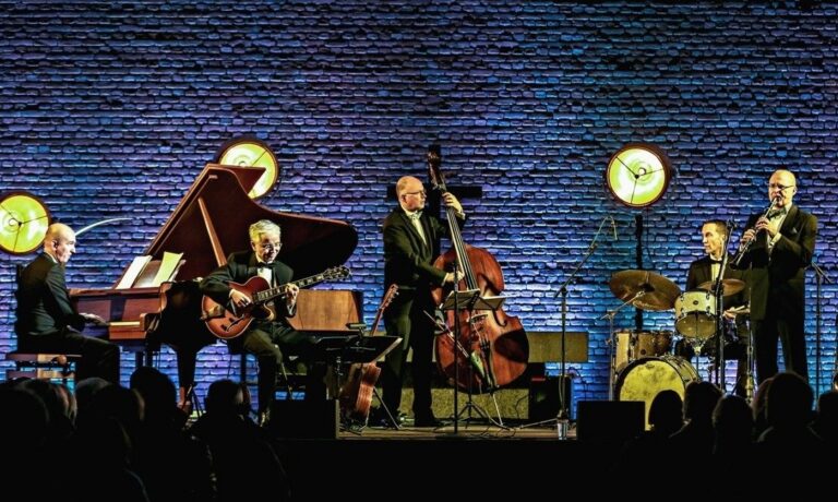 Fünf Musiker in Anzügen spielen Jazz auf einer Bühne mit Klavier, Gitarre, Kontrabass, Schlagzeug und Klarinette vor
