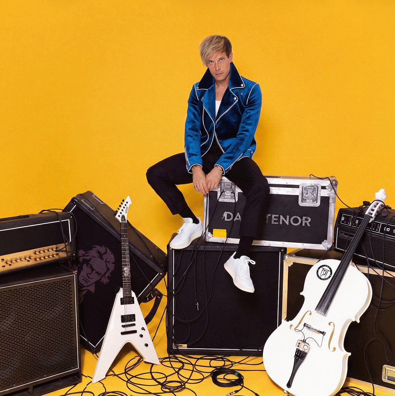 The Dark Tenor sitzt auf Verstärkern und Roadcases umgeben von einer weißen E-Gitarre und einem weißen Cello vor gelbem