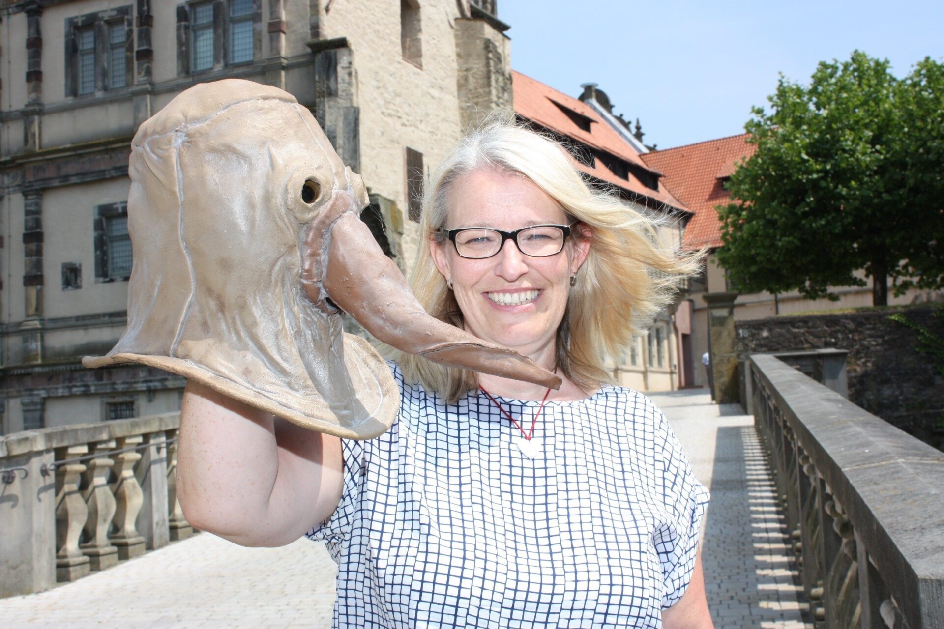Dr. Susanne Hilker lächelt vor einem Schloss und hält eine beigefarbene Pestdoktor-Maske mit Schnabel