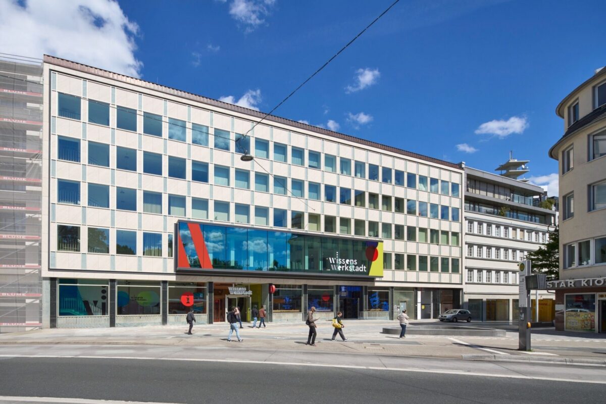 Menschen gehen auf dem Bürgersteig vor dem Gebäude WissensWerkstadt unter blauem Himmel mit einigen Wolken