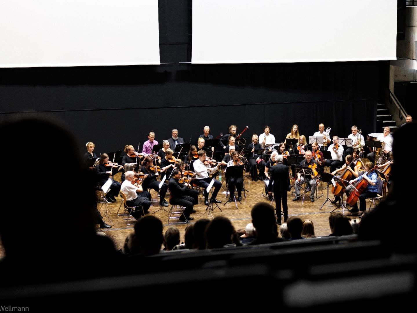 Orchester mit vielen Musikern und einem Dirigenten probt auf einer Bühne vor Publikum
