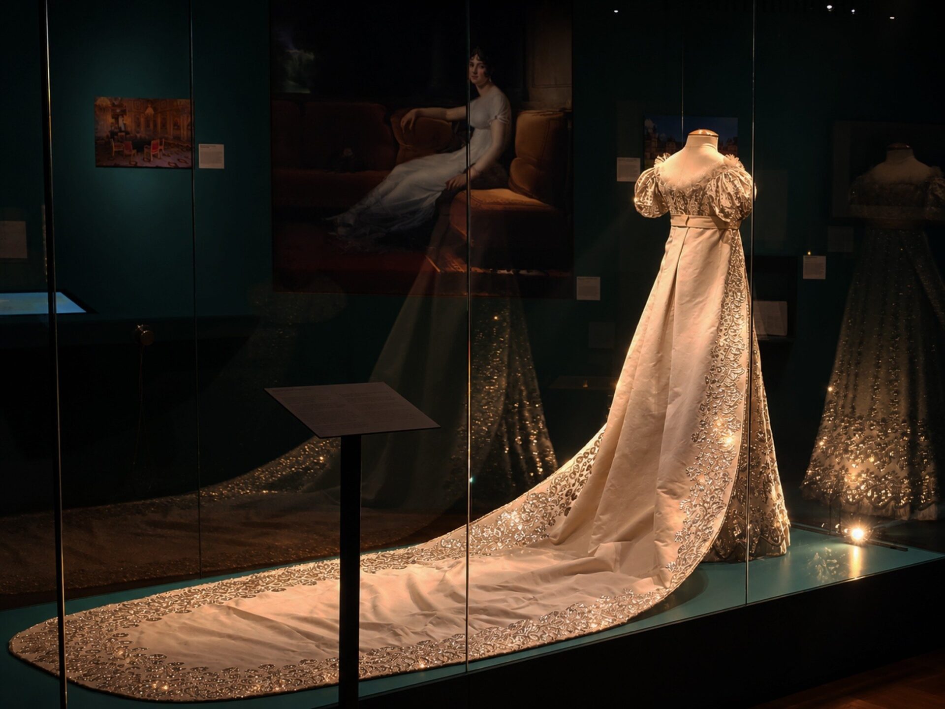 Ein helles, langes Kleid mit goldener Stickerei und Schleppe auf einer Puppe in einer beleuchteten Museumsvitrine,
