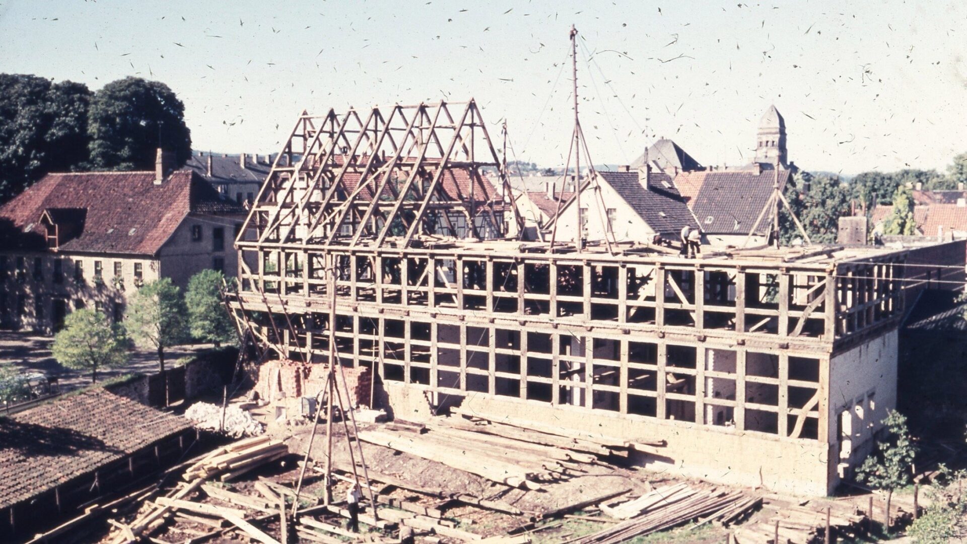 Großer Fachwerkbau im Rohbau mit sichtbarem Holzgerüst und Bauarbeitern, umgeben von Häusern und Bäumen