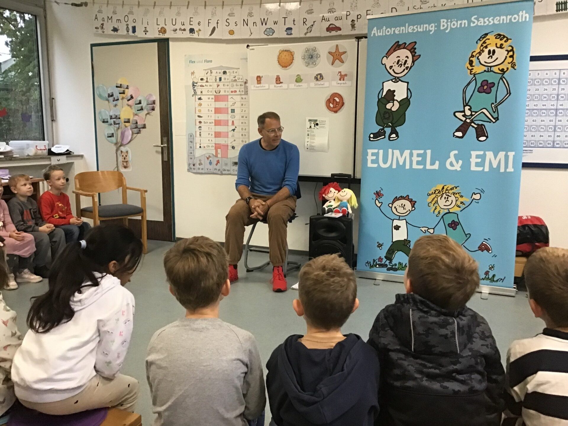 Autor Björn Sassenroth liest Erstklässlern in einem Klassenzimmer vor, neben ihm ein Banner der Figuren Eumel und Emi