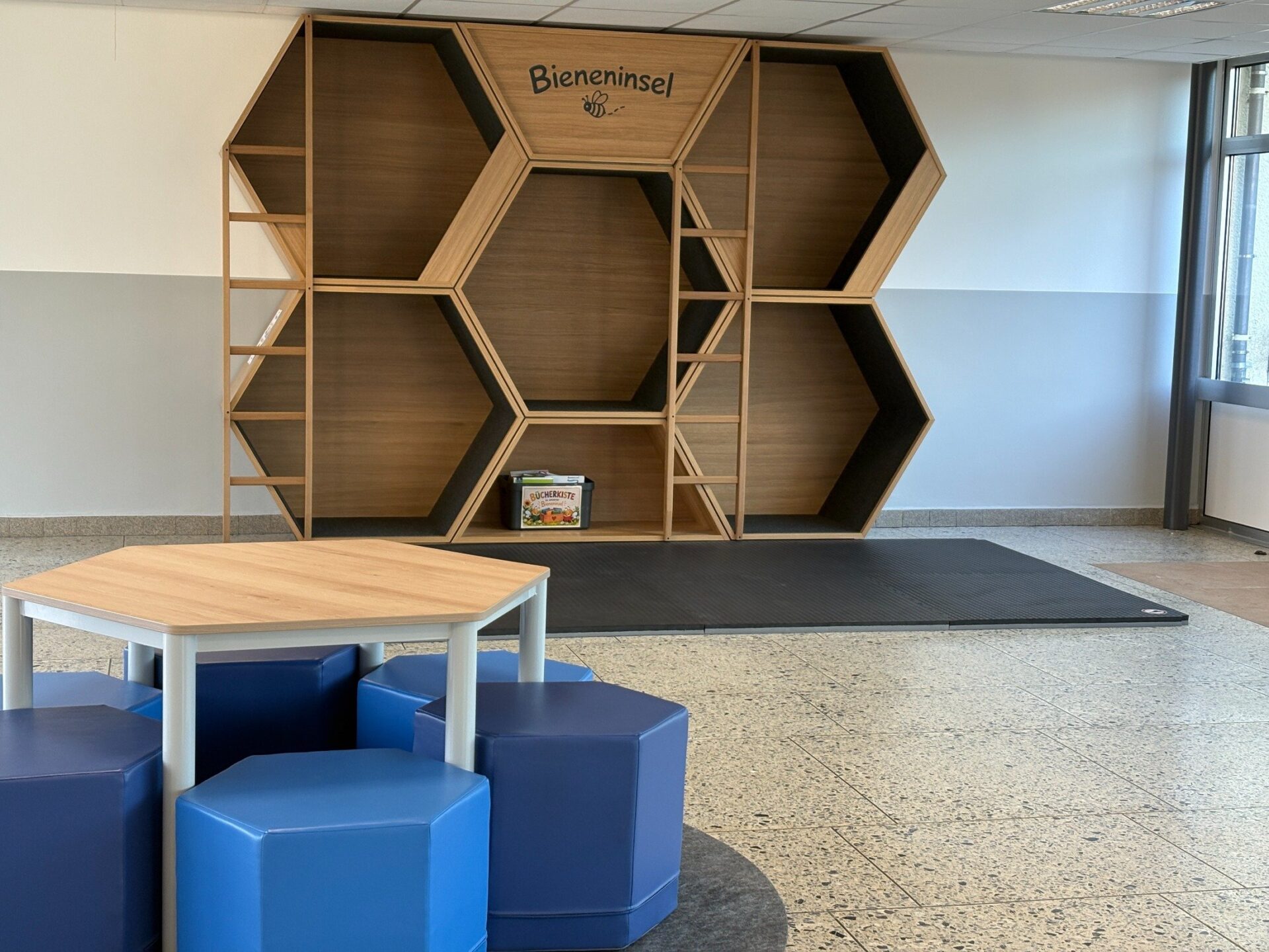 Ein Wabenregal mit der Aufschrift „Bieneninsel“, einer Bücherkiste und davor ein sechseckiger Holztisch mit blauen