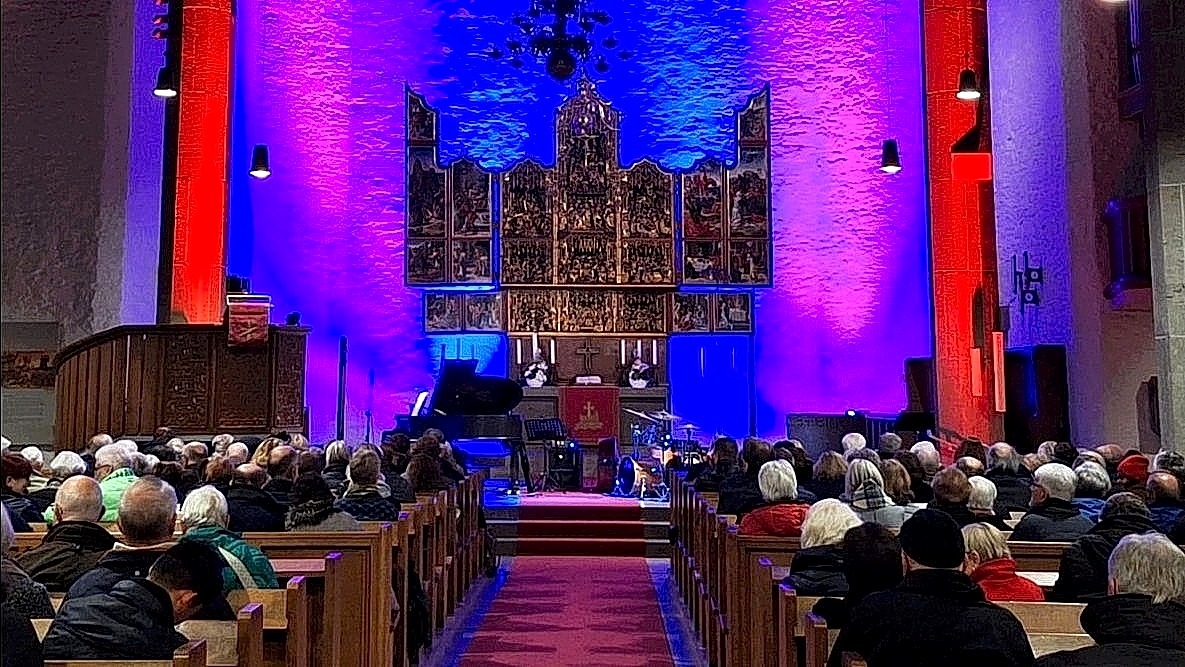 Konzert in einer Kirche: Publikum in Holzbänken, Bühne mit Flügel und Schlagzeug vor einem blau und rot beleuchteten