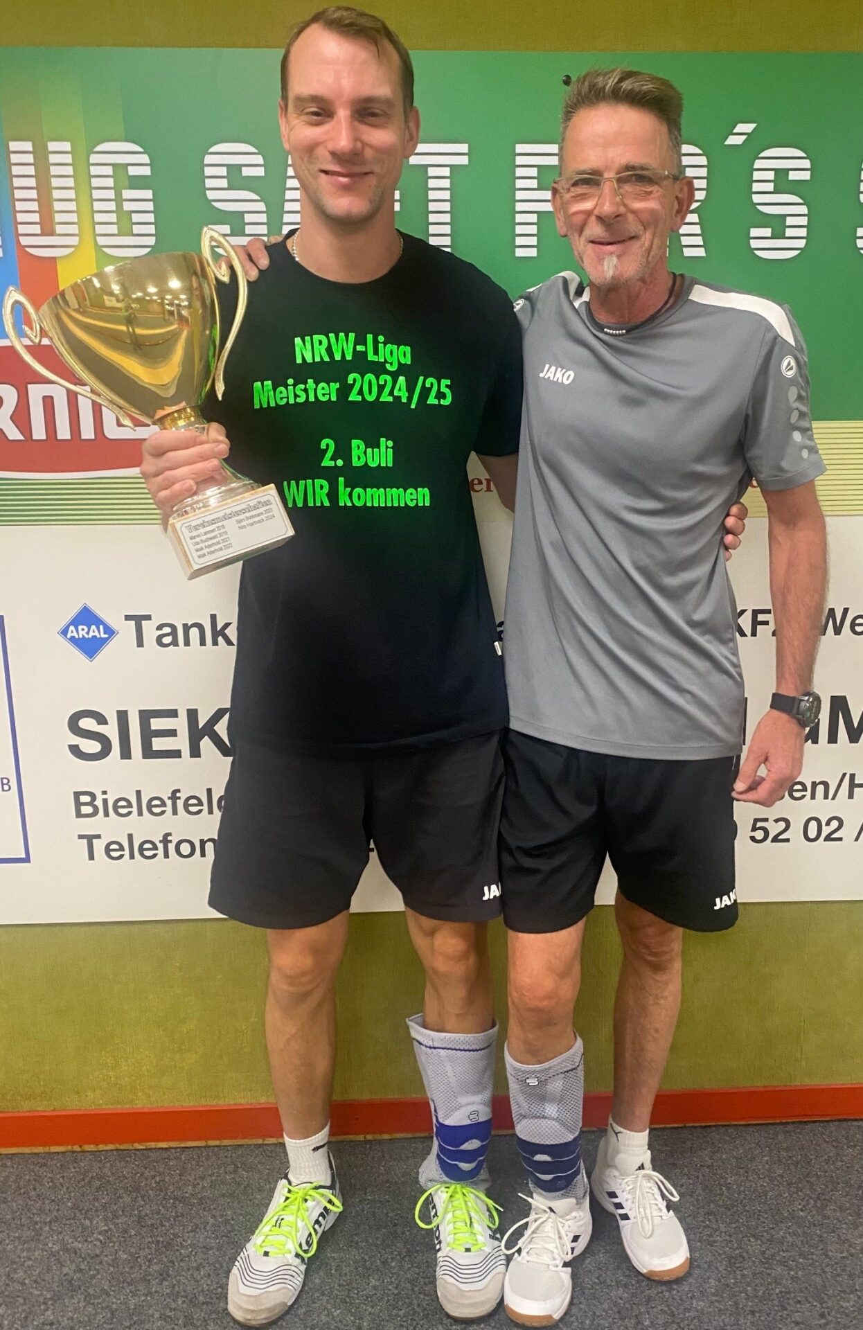 Nils Hartnack hält strahlend einen goldenen Pokal und steht Arm in Arm mit einem anderen lächelnden Mann