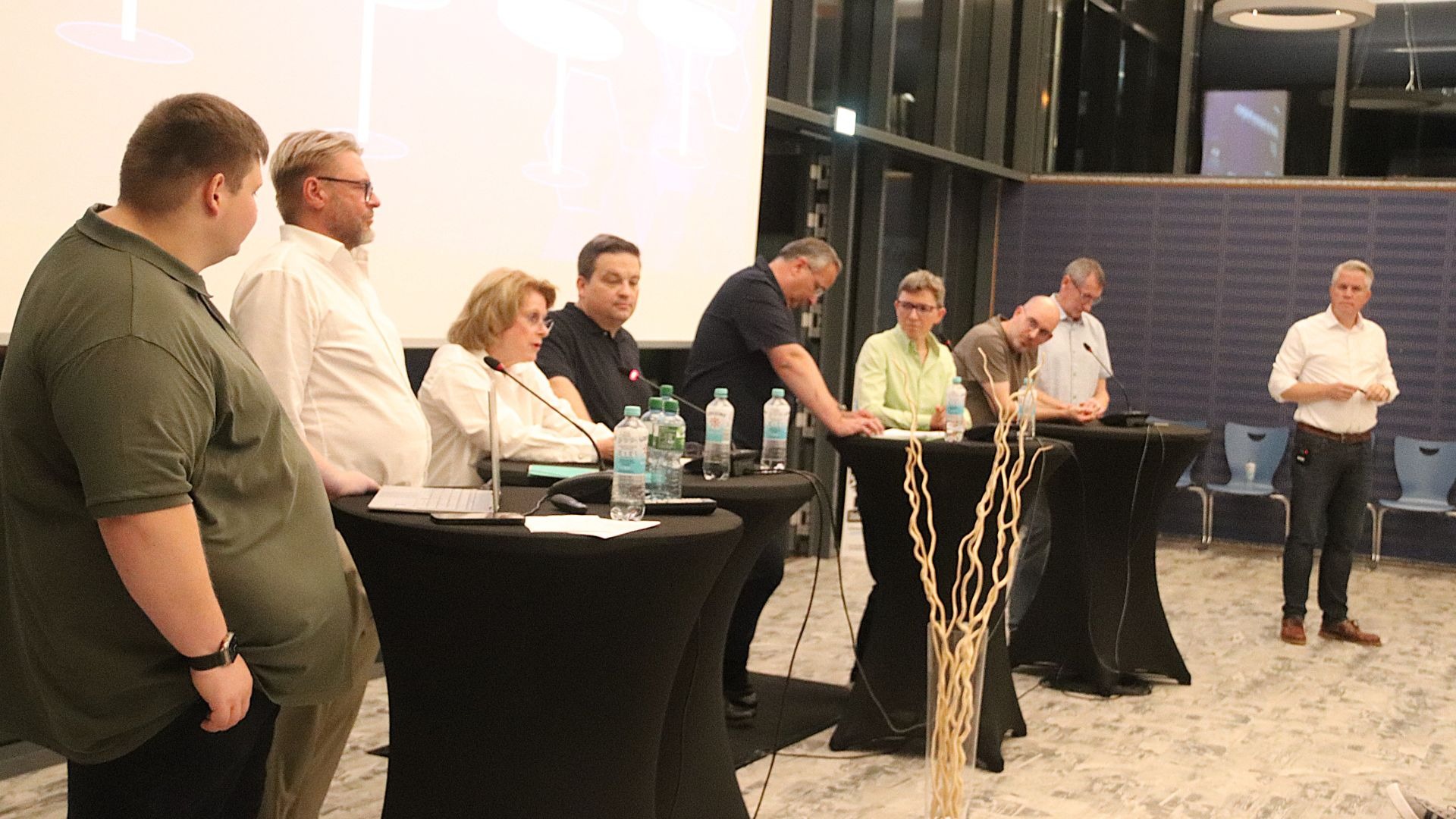 Neun Erwachsene diskutieren an Stehtischen mit Mikrofonen und Laptops vor einer großen Projektionsfläche in einem Raum