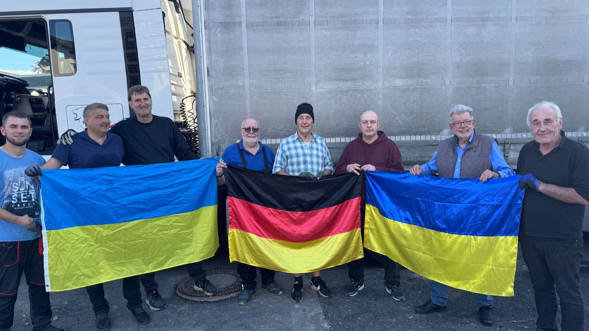 Acht Männer stehen vor einem Lastwagen und halten zwei ukrainische und eine deutsche Flagge