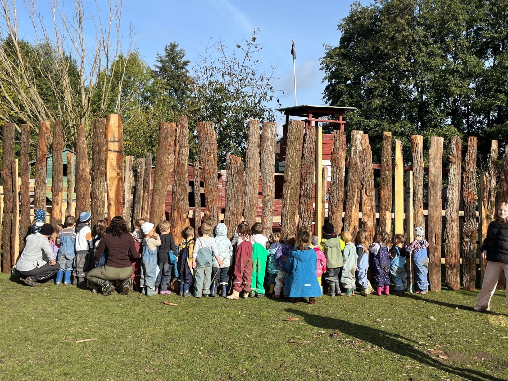 Kinder und Erwachsene stehen an einem Holzstammzaun und blicken auf ein rotes Holzhaus mit schwarzer Flagge